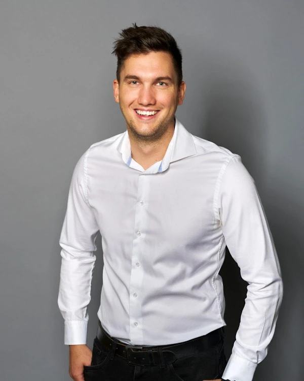 Patrick Haidacher; -	Der Mitarbeiter für die Technische Leitung am Standort in Österreich trägt ein weißes Hemd, hat seine Haltung leicht nach vorne gebeugt und lacht mit breitem Mund in die Kamera.
